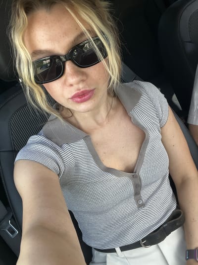 A woman with blonde hair is pictured indoors, possibly in a car, as part of a selfie-style photograph. She wears dark sunglasses and has pink lipstick on. She's dressed in a gray and white striped top with buttons, and a dark brown belt is visible. A smartwatch is on her left wrist. The background includes dark car seats and white clothing. The location details (city of Bobrov, Russia) cannot be confirmed from the image.