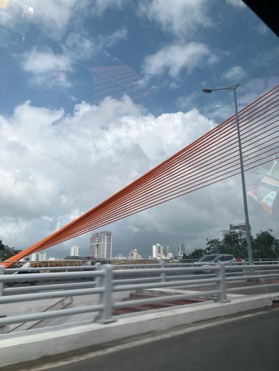 The image depicts a daytime scene from the Trần Thị Lý Bridge, looking generally east or southeast towards the city skyline, located in Da Nang, Vietnam, with elements of Hòa Cường Ward visible or implied nearby. The sky features a mix of clear blue patches and large, white cumulus clouds, with darker, more overcast clouds on the horizon.

In the foreground, the asphalt road surface of the bridge is visible, with a light gray concrete barrier and multi-bar metal guardrail running along the right side. Several parallel orange cable stays of the bridge ascend diagonally from the lower-left to the upper-right of the frame. A tall gray street light pole with a curved lamp arm stands on the right side, extending upwards. Reflections of the vehicle's interior and external objects are visible in the upper portion of the image, suggesting the photograph was taken through a window.

On the bridge road, a dark-colored SUV is partially visible, and other vehicles appear as blurs further along. Beyond the bridge's immediate structure, a cityscape is visible, dominated by numerous multi-story buildings and high-rise structures, including a prominent, light-colored tall building with distinct vertical elements. Further in the background, another bridge structure with yellow/brown trusswork is discernible. Sparse green foliage is also present along the distant shoreline, bordering some of the lower buildings.