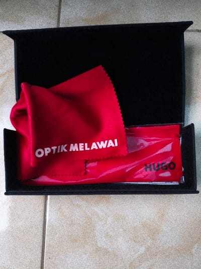 The scene, located in Bojongherang, Indonesia, depicts an open, black, rectangular hard-shell case resting on a light-colored tiled floor. Inside the case, on the left, is a folded red microfiber cleaning cloth displaying the white uppercase text "OPTIK MELAWAI". To the right of the cleaning cloth, within the same case, lies a red, slightly translucent synthetic pouch or bag. This pouch bears the black uppercase text "HUGO" on its visible surface. No individuals are present. The tiled floor exhibits light-colored, possibly beige or off-white, square tiles with visible grout lines, suggesting an indoor setting.