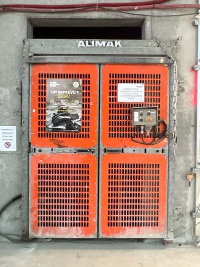 The image displays an ALIMAK industrial construction hoist door, positioned within a concrete structure, likely at a construction site in Aulnay-sous-Bois, France. The door assembly features a grey metal frame enclosing four orange perforated panels arranged in a 2x2 grid, with perforations formed by vertical slots. Above the frame, a dark, open rectangular shaft entrance is visible, with red corrugated conduit pipes running horizontally overhead. The door and surrounding concrete show signs of dust and usage. On the upper left orange panel, a laminated poster in French, titled "UN IMPRÉVU? STOP!", details a three-step incident response procedure and displays the "Société des Grands Projets" logo. The upper right orange panel bears a white sign in French, stating, "PLEASE GO TO THE SECURITY POST FOR THE OPENING OF THIS DOOR." A grey control panel with black, green, and red (emergency stop) buttons, connected by black wires, is mounted on this same panel. A metal chain hangs from the upper right of the door frame, and a yellow padlock is visible on a bracket attached to the right concrete wall. A circular white prohibition sign on the adjacent left concrete wall, also in French, reads, "PROHIBITION TO USE THE LIFT IN CASE OF FIRE." No individuals are present in the frame.