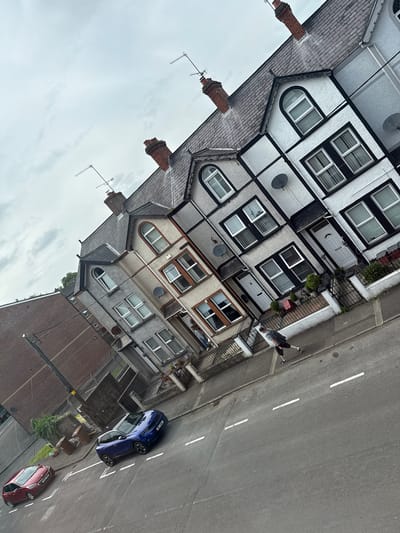 This image captures a street scene in Ballymena, United Kingdom. The perspective is from an elevated viewpoint, looking down at a row of terraced houses and the street below. The sky is overcast with clouds.
In the foreground, a grey asphalt road with white dashed lines marking lanes is visible. Two cars are parked on the street: a red hatchback on the left and a blue SUV on the right. A single person, wearing a grey t-shirt, black shorts, and a backpack, walks along the pavement on the right side of the road. The pavement is bordered by a low metal fence and the front gardens of the terraced houses.
The terraced houses are constructed in a style typical of the United Kingdom, with pitched roofs covered in grey tiles and brick or rendered facades. Some of the houses have black-and-white contrasting detailing around the windows and rooflines. Many windows are visible, some with white blinds or curtains. Chimneys extend from the roofs, topped with red brick. Satellite dishes are attached to the facades of some of the houses. A large brick building with a sloped roof is situated to the left of the terraced houses.
The street appears to be residential, with a general sense of urban housing. There are some trees and plants visible in the gardens and along the street. The overall impression is of a moderately populated street on a cloudy day.