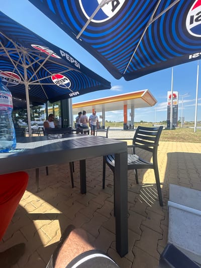 The image captures a sunny outdoor seating area with a gas station in the background. Two large umbrellas with the Pepsi logo provide shade over a table and chairs. On the table, a large bottle of water and an ashtray are visible. A man wearing a white tank top and sunglasses is seated at the table, facing left. In the background, two women and another man are standing near a building. The gas station has a distinctive orange and white canopy, and a sign displays fuel prices. The ground is paved with light brown interlocking bricks. The sky is clear and blue. The scene appears to be a rest stop or service area on a road, possibly near Polsko Padarevo, Bulgaria, given the presence of the "rom petrol" sign which indicates a Bulgarian petrol station chain.