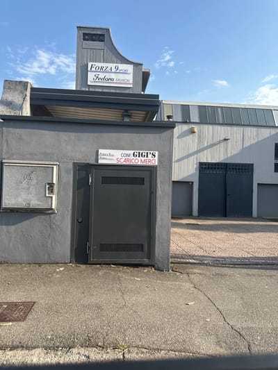 The image shows an exterior view of a commercial building with two signs. The larger sign, mounted on a grey concrete structure, reads "FORZA 9 SPORT Fedora FASHION" in a stylized font. Below this, a smaller white sign on a grey wall displays "FORZA 9 SPORT Fedora FASHION". To the right of this sign is another white sign with black text that reads "CONF. GIGI'S SCARICO MERCI". Adjacent to the signs on the left is a grey utility box with "ENEL" printed on it. A dark grey metal door with a gridded section is visible to the right of the utility box. The building's exterior is predominantly grey concrete and white panels, with a large set of dark grey double doors in the background. The ground in front of the building is paved with patterned tiles. The sky above is blue with scattered white clouds. This scene is located in Rudiano, Italy.