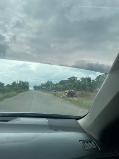 This is a vertical, slightly tilted, eye-level, first-person perspective photograph taken from inside a vehicle, looking out the side window. The scene depicts a rural road in Junyura, Nigeria, under a cloudy sky.

The left side of the image is dominated by dark, heavy clouds, suggesting impending rain or overcast conditions. The right side of the window reveals a dusty, unpaved road stretching into the distance. Flanking the road are lush green trees, including what appear to be banana plants, and dense foliage. In the foreground on the right side of the road, there is a small pile of stacked wood. Further down the road, a lone motorcycle can be seen. Electrical poles with visible wires run along the side of the road.

The interior of the vehicle is partially visible in the upper right corner, showing a grey plastic dashboard with an air vent and a speaker grille. The overall impression is one of traveling through a tropical, rural landscape on a potentially humid or rainy day. The light suggests it could be mid-morning or afternoon, but the heavy cloud cover obscures direct sunlight. There is no visible text in the image.