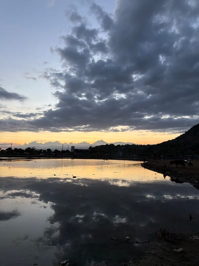 The image shows a waterfront scene in Upt Tambak Sari, Indonesia. On the left, dark, billowing clouds dominate the sky. The horizon is illuminated by a warm, yellow light that appears to be either sunrise or sunset. In the center, a distant mountain range is visible, its silhouette punctuated by a single prominent peak. Closer to the viewer, there are buildings and utility poles lining the waterfront. The right side of the image features a murky body of water which seems to reflect the clouds and golden sky, creating a mirrored effect. Along the shoreline, debris and trash are visible. The scene conveys a sense of environmental neglect juxtaposed with the natural beauty of the sky and mountains. No people are visible.