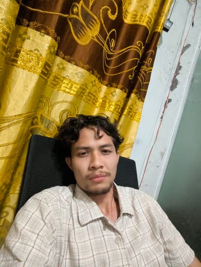 In Pulodolang, Indonesia, a young man with curly dark hair and a mustache is seated in a black chair, facing forward and looking at the camera. He is wearing a light-colored plaid shirt. Behind him, a draped curtain features a bold, ornate pattern in shades of brown and gold, with large floral motifs and swirling lines. To the right of the curtain, a white wall is visible, with a red wire running vertically down its surface. The overall scene suggests an indoor setting, possibly a room within a residence.