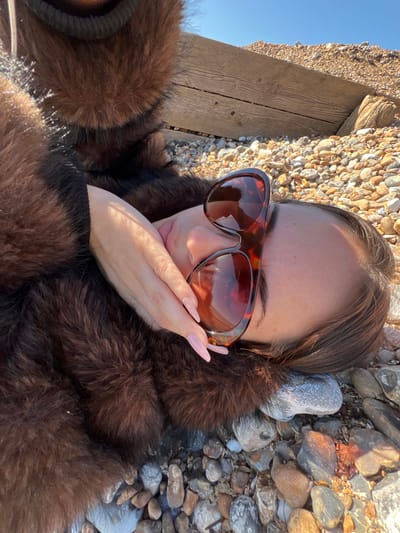 The image presents an overhead, close-up view of an individual lying supine on a pebble beach in Worthing, United Kingdom. The subject's upper torso and head are central, adorned in a thick, dark brown, possibly faux fur, garment featuring a dark ribbed collar. Large, tortoiseshell-patterned sunglasses obscure their eyes and nose. A hand with pale pink, manicured fingernails rests adjacent to the left side of the face, near the sunglasses. Dark brown hair frames the face, partially visible around the edges of the sunglasses and against the pebbles. The ground consists of numerous smooth, multi-colored pebbles. In the background, weathered wooden planks, consistent with a groyne or sea defense, extend horizontally. Above the planks, a clear, bright blue sky is visible. The scene is illuminated by strong, natural sunlight, casting subtle highlights and reflections, particularly on the sunglasses.