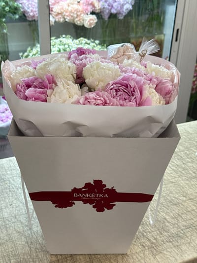 A bouquet of pink and white peonies is presented in a light gray gift bag. The flowers are wrapped in pale pink paper, with a clear plastic covering secured by twine at the top. The gift bag has a dark red, stylized floral design and text that reads "BANKETKA Flower Boutique". In the background, through a window or glass partition, other floral arrangements in various shades of pink and purple can be seen. The setting appears to be a flower shop or a similar retail environment, indicated by the display of flowers and the branding on the bag. The lighting suggests daytime. The image focuses on the presentation of the floral gift.