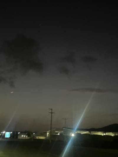 The image depicts a dim, twilight sky with some clouds. A crescent moon is visible in the upper left portion of the sky. In the middle ground, there are several structures including a utility pole with crossbars, and what appear to be industrial or commercial buildings. Artificial lights are present, illuminating some of the buildings and creating lens flares in the image. A faint mountainous or hilly silhouette is visible on the horizon in the background. The foreground shows a dark, unlit area, possibly a road or ground. There are no people or visible actions in the scene. The overall impression is of an industrial or semi-urban area at dusk or night. There is no specific information in the image to confirm the location as Conuco Viejo, Venezuela.