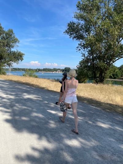 The image depicts two individuals walking along a gravel path next to a body of water. The person in front is wearing a straw hat, a light pink tank top, and white shorts. They are barefoot and appear to be reaching for something behind them. The second person, partially obscured, is wearing a black tank top and patterned shorts. They are also barefoot. The scene suggests a sunny day, with clear blue skies and dappled shadows from trees on the path. The background features dry, grassy terrain, trees, and a distant shoreline with more trees and some structures. Boats and people can be seen on the water, indicating a recreational area. The overall atmosphere appears relaxed and leisurely, consistent with a warm day outdoors in a natural setting. There is no visible text in the image.