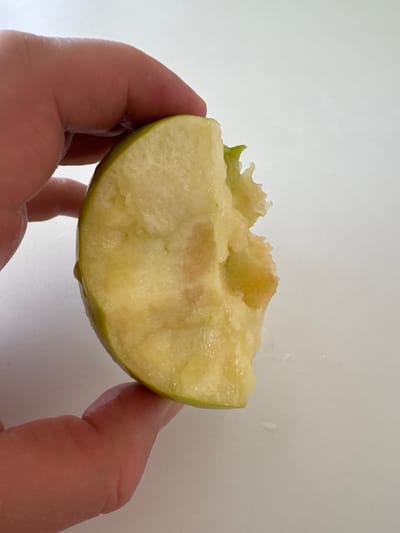 A close-up shot captures a person's hand holding a wedge of a green apple. The apple appears to have been bitten into, revealing a pale yellow, juicy interior with some brown discoloration. A small droplet of juice is visible hanging from the bottom edge of the apple. The background is a plain, white surface, suggesting an indoor setting. The lighting is bright and even, indicating it could be daytime with artificial light. The image focuses on the texture and freshness of the apple, with the hand providing a sense of scale and human interaction with the fruit. There is no visible text in the image.
