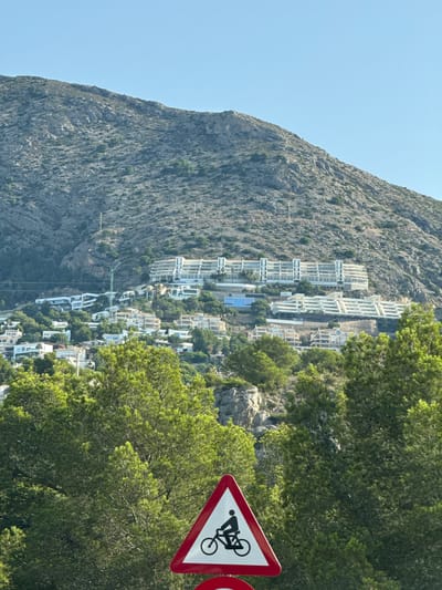 This is a medium shot, eye-level, vertical photograph taken on a sunny day, featuring a hillside populated with buildings and lush green trees, with a triangular warning sign in the foreground. The sign, edged in red and white, displays a black silhouette of a person riding a bicycle, indicating a cycling hazard or route. Behind the sign, dense green trees fill the lower half of the frame. The middle ground is occupied by numerous white buildings, varying in size and architectural style, clinging to the hillside. A prominent, large, white multi-story building with many balconies stands out on the upper part of the developed slope. Further up the hillside, rocky terrain with sparse vegetation extends to the top of the frame, where a clear, bright blue sky dominates. A green construction crane is visible among the buildings on the hillside. This scene is characteristic of the coastal areas around Altea, Spain, known for their terraced developments on steep slopes overlooking the Mediterranean.