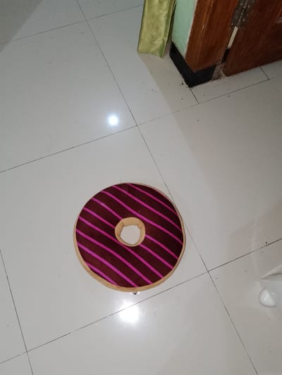 In Sendangrejo, Indonesia, a brown donut-shaped cushion with pink stripes lies on a white tiled floor. The cushion is centrally located in the frame. The floor is clean and features visible grout lines. In the background, a wooden door frame and a portion of a wall covered in green fabric are visible. A bright light reflection is present on the floor.