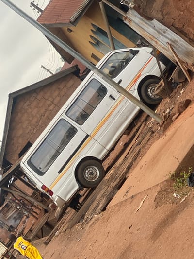 The image depicts an outdoor scene in Okura, Nigeria, presented at a diagonal angle. A white passenger minivan, featuring yellow and orange horizontal stripes along its lower body, is stationary on an elevated reddish-brown dirt embankment. The driver's side of the vehicle is visible. Behind the van are two buildings: one is a light yellow/ochre structure with a red corrugated metal roof, characterized by distinctive arched gable ornamentation, possibly indicating a church; to its immediate left is another building constructed from reddish-brown unfired bricks or concrete blocks, also with a corrugated metal roof. A slender silver-colored metal pole extends diagonally across the frame, partially obscuring the van's front section and portions of the buildings. The ground consists of reddish-brown laterite soil, with scattered wooden planks, corrugated metal sheets, and miscellaneous debris visible near the structures and the van. In the bottom-left foreground, a person, likely a child or youth, is seen from behind, wearing a yellow sports jersey bearing the name "JAMES" and the number "6." A dark-colored dog is standing directly beneath the rear section of the white minivan. The sky overhead appears overcast or hazy grey, indicating daytime conditions.