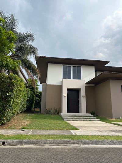 The image shows the front of a house with a brown door, a concrete walkway, and a concrete porch with steps leading to the door. The house has a white exterior and a brown roof. It is located at coordinates 9.945, -84.201 in Rio Oro, Costa Rica, and sits amidst lush green foliage. To the left of the home is a large, green bush and a palm tree. There are light fixtures on the front of the home near the door and a small plant to the right of the steps. The sky is overcast with white clouds.