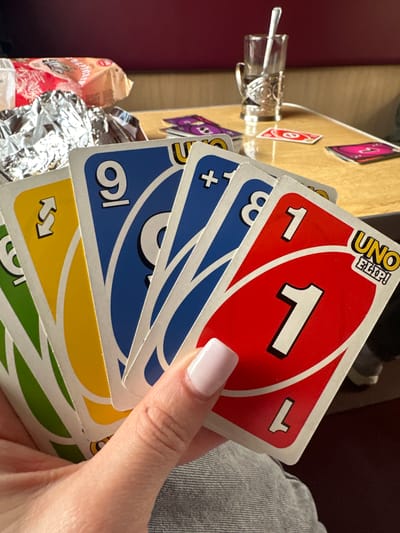 A hand holds a stack of Uno Flip! cards. The top card is red with a white "1". Below it are several blue cards, one with "3" and another with a "+" symbol. Further down are a blue card with "6", a yellow reverse card, and a green card. The cards are held over a cluttered table with a clear glass object, partially unwrapped food packaging, and a card holder with cards in it. The background includes a grey fabric and part of a red surface. The location is likely indoors in Мастоўскі сельскі Савет, Belarus.