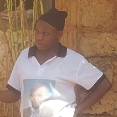 An individual is depicted standing outdoors in the city of Haifam, Nigeria. The subject, a person with dark skin, wears a black skullcap and a white short-sleeved polo shirt with a black collar and black trim on the sleeve ends. The front of the shirt displays a printed image of a light-skinned male figure, possibly an adult, wearing a suit jacket and collared shirt against a light blue background. The individual looks towards the left side of the frame with a neutral expression. Their left hand is positioned on their hip, while their right arm is partially visible, bent at the elbow. The background on the left features vertical, light-colored plant stalks. To the right of the subject, a wall constructed from irregular, light brown stone or brick is visible. The scene is brightly illuminated by natural daylight.