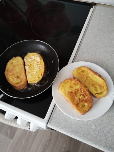 The image shows two pieces of French toast being cooked in a black frying pan on a stovetop. One piece of French toast is being fried in oil, and the other piece has grated cheese sprinkled on top. To the right of the frying pan is a white plate with two cooked pieces of French toast. The French toast appears golden brown and slightly crispy. The setting is a kitchen, with the stovetop and countertop visible. The lighting suggests it is daytime. There are no people visible in the image. The image does not contain any visible text.