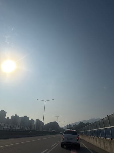 The image depicts a view from a vehicle traveling on a highway, likely in Namyangju-si, South Korea. The sun is bright and high in the sky, casting a glare that illuminates the scene. On the left side of the highway, a row of apartment buildings can be seen, some of which appear to be under construction or renovation. A few streetlights line the road.

In the foreground, a white SUV is driving on the highway. Its license plate is visible, reading "396가 4940". Other vehicles are also present on the highway, though less prominently. On the right side of the highway, a sound barrier wall lines the road, with a view of hills or mountains in the background. The sky is clear with a faint haze.