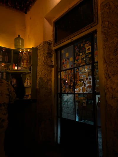 This image depicts a dimly lit, warm interior scene, likely a bar or a cozy establishment. The overall atmosphere is dark and intimate, illuminated by localized artificial lighting.

A significant feature is a large window or glass-paned door in the upper center, completely covered with a dense collection of diverse stickers. Through the lower right section of this window, a very dark, unidentifiable exterior suggests nighttime.

On the right side of the frame, a rustic wooden shelving unit is visible. It holds several bottles, small decorative string lights, and a religious icon or framed picture, possibly of the Virgin Mary, also adorned with lights. A person is partially visible below this, seated or leaning, with light subtly illuminating their face and glasses as they look downwards, possibly at a phone. They are wearing a patterned shirt.

To the left of the shelving unit, a large, translucent green bottle or carboy rests on a surface. The walls and ceiling have a rough, textured finish, possibly stone or plaster, painted in warm, earthy tones. On the far left edge, a stack of what appears to be wooden planks or wine bottles is visible.

Visible text from the numerous stickers includes "NO BAD DAYS" (repeated several times), "R", "KEEPTHELINE", "Steiermark", "MALEX", "Jew", and "Jefe". The rustic decor, the religious icon, and the warm, low lighting suggest a character-filled venue, possibly a bar or café in Mérida, Mexico, providing a relaxed and inviting setting for an evening gathering.