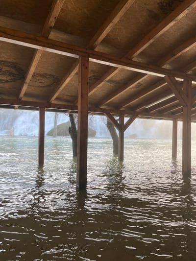 The image displays an outdoor scene at Kravica Waterfalls in Zvirići, Bosnia and Herzegovina. The foreground features a wooden, open-sided canopy structure with a sloped roof composed of exposed wooden beams and panels, likely oriented strand board (OSB). This structure is supported by multiple square-sectioned wooden posts extending vertically into a body of dark, rippled water. The water level is elevated, submerging the lower portions of the support posts. Beyond the structure, partially submerged in the water, are several bare tree trunks and a large, rounded rock formation. In the distant background, a wide, multi-tiered waterfall is visible, generating a significant mist that diffuses the ambient light. No individuals or specific activities are discernible within the frame.