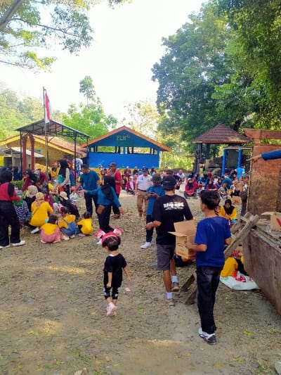 The image depicts an outdoor gathering of numerous individuals, including adults and children, on unpaved ground in Jetis, Indonesia. In the background, to the left, a small building with a red and white Indonesian flag on a pole is visible. Adjacent to it, a blue building with an orange roof displays the white text "PLN Peduli." To the right of the blue building, a brown-roofed, open-sided gazebo structure stands.
Groups of people are seated on the ground, some on a white tarp, with several individuals wearing yellow or other brightly colored clothing. Other adults are standing and interacting. In the mid-foreground, a male individual in a black t-shirt and grey shorts holds a cigarette, while another male in a blue t-shirt and dark pants carries a brown cardboard box. A small child in a black outfit and pink shoes, with red and white hair accessories, walks towards the lower center of the frame. Some children are observed holding small Indonesian flags. The scene indicates a community event, possibly involving distribution, organized by PLN Peduli.