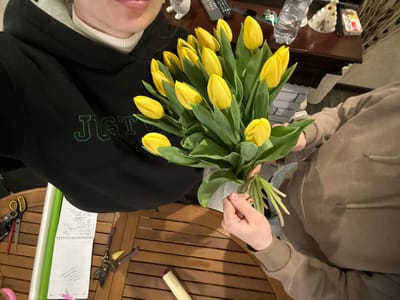 The image captures an indoor scene in what is identified as Slavyansk-na-Kubani, Russia, focusing on a bouquet of yellow tulips and two partially visible individuals.
In the foreground, a slatted light brown wooden table is visible. On the table's left side, various floral arrangement tools are laid out: a pair of gold-handled scissors, a pair of red-handled scissors, a roll of white or light green floral wrapping material, an open white notebook with handwritten text, a pair of purple-handled gardening shears, a small yellow object, and a pink ribbon roll. A light-colored cylindrical object rests further right on the table.
Two individuals are present. On the left, an individual's upper torso and lower face (lips and chin) are visible. This person wears a black hoodie with a grey inner lining and a white turtleneck sweater underneath. The black hoodie features green embroidered text on the chest, partially obscured, appearing to begin with "JUST." On the right, a second individual's arms and upper torso are visible. This person wears a light brown or beige long-sleeved top and is holding a bouquet of approximately 15-20 bright yellow tulips by their green stems, which are tied together with a thin, light-colored string. The stems below the tie appear recently trimmed.
In the background, behind the individuals and above the table, a dark brown polished wooden surface, possibly a shelf or dresser, is visible. Items on this surface include a dark remote control, a plastic water bottle, a cluster of keys, a white decorative ceramic figurine, and a small white box with red lettering, identifiable as "CLUTCH" cigarettes. The scene is illuminated by artificial indoor lighting.