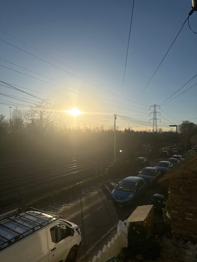 The image captures an elevated view of an urban landscape during either sunrise or sunset, looking generally eastward from a residential area in St Denys, Southampton, United Kingdom.
The upper portion of the frame features a clear, pale blue sky with a prominent, bright sun positioned in the upper-center left, creating a significant lens flare and backlighting effect. Numerous thin overhead power lines and communication cables crisscross the sky. A large, lattice-structured electrical transmission pylon stands in the distant midground, to the right of the sun's glare.
The midground is dominated by a multi-track railway line, featuring at least four parallel tracks running horizontally across the left side of the frame. A security fence separates the railway from an adjacent road. Beyond the tracks, a dense line of bare deciduous trees and shrubs is silhouetted against the bright sun. A utility pole with multiple attached cables is visible between the railway and the street.
In the foreground and extending into the midground on the right, a residential street, likely Northam Road, is visible. The street surface is dark asphalt, illuminated by the low sun angle, which casts long, distinct parallel shadows across it. A white commercial van with a roof rack is parked on the left side of the street, partially in shadow. Further along the street, a dark blue hatchback car (with registration WG59 KSS) is prominently parked, facing towards the viewer. A line of other parked vehicles extends down the street into the distance. On the immediate right, a section of a low, white, scalloped wall or fence from a residential property is visible, with dark foliage behind it. No individuals are present within the scene.