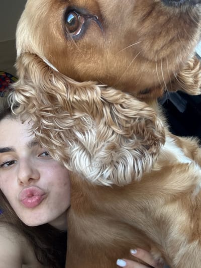A close-up, selfie-style portrait shows a young woman with brown hair, her lips pursed in a kiss, and a large, golden-brown dog positioned in the foreground. The dog's head is partially obscured, but its large, brown eye is visible, reflecting light. The woman's left eye, eyebrows, and nose are also visible, with her skin appearing slightly flushed. The dog's long, wavy fur is prominent, with some lighter strands mixed in. The background is indistinct and appears to be indoors, with a hint of a colorful object on the left. There is no discernible indication of the location being Dibich, Bulgaria.