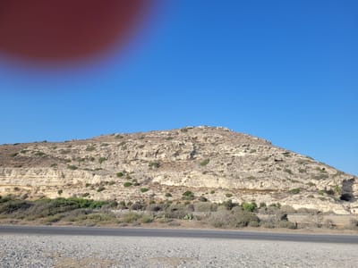 The image shows a large, light-colored, rocky hill with sparse vegetation on its slopes. The hill has layered sedimentary rock formations and some caves or hollows are visible. At the base of the hill, there is a road in the foreground, with a gravel shoulder and a paved surface. Behind the road, there is an area of dense scrub and small trees. The sky is clear and bright blue. The scene is outdoors, under natural daylight. There are no people visible in the image. The location is identified as Επισκοπή Λεμεσού, Cyprus. A large reddish-brown blur, likely a finger or thumb, partially obscures the top left corner of the image.