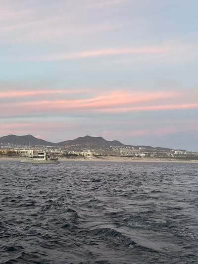 The image depicts a view of Cabo San Lucas, Mexico, from the sea. On the right is dark, choppy water. In the center is the coastline with buildings and mountains. The sky on the left exhibits a light color gradient with pink and orange streaks.