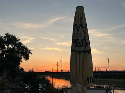 The image depicts a sunset scene over the Rhine River in Mannheim, Germany. In the foreground, a closed patio umbrella, cream-colored with black lettering and a decorative pattern, stands prominently on the right side. The umbrella is tied with a white strap. To the left, a large, silhouetted tree with dense foliage is visible.
In the midground, a bridge spans the river, with a road or railway line on top. Several figures, appearing as small silhouettes, are walking across the bridge. A car is also visible on the bridge. The river reflects the warm hues of the sunset, creating a shimmering orange and yellow surface. Along the riverbank, there are trees and some structures.
The sky is painted with the soft oranges and pale yellows of sunset, with wispy clouds scattered across it. The overall atmosphere is tranquil and scenic, characteristic of an evening by the river.