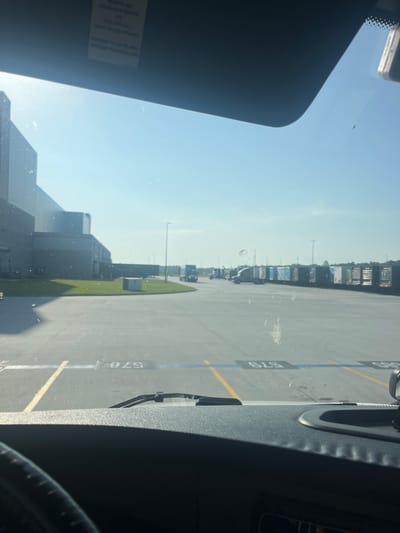 The image captures a view from inside a truck cab, looking out through the windshield on a bright, sunny day. The scene is an outdoor industrial area, likely a logistics hub or distribution center. Dominating the left side of the frame is a large, modern building with a grey facade, typical of a warehouse. In front of the building, a patch of green grass is visible.
The majority of the image is taken up by a vast, paved lot, marked with yellow and white lines indicating parking or driving lanes. Scattered across this lot are numerous semi-trailer trucks, parked in neat rows. Many of these trailers are white or a light color, with some featuring blue accents and branding that suggests Amazon. The trucks are stationary, indicating a period of rest or waiting.
In the distance, more trucks and buildings can be seen, suggesting a large-scale operation. The sky is clear and blue, with a bright sun casting shadows and creating a glare on the windshield, which also shows some dirt and a spiderweb. The foreground shows the dashboard and steering wheel of the truck, offering a sense of the viewer's perspective. The number "578" is visible on the pavement in front of the truck, likely a parking spot or bay number. The location is identified as Pooler, United States of America.