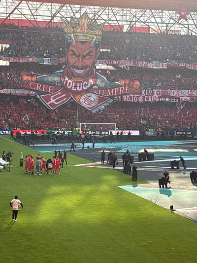 The image shows a stadium filled with fans, many holding red and white banners and signs, cheering for the Toluca Deportivo football team. A large banner depicting a crowned devil, the team's mascot, dominates the scene. Players in red uniforms are lined up on the green field. Security personnel in black uniforms are positioned along the edge of the field. The environment includes stadium architecture and advertisements for Hilton and Riyadh Air. The location is likely in Toluca, Mexico, the home city of the Toluca Deportivo team.