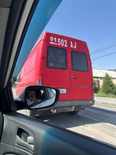The image captures a view from inside a car, looking out at a red van. The van's rear doors are visible, with large white lettering that reads "01502 AJ KG" on the left door and "2 AJ" on a license plate below the right door. The word "Sprinter" is visible in smaller white text on the right side of the van's rear.

A side-view mirror of the car reflects the interior of the car and a person holding a smartphone, presumably taking the photo. The person in the mirror is looking at the phone.

The scene appears to be a roadside setting, with a paved road and a gravel shoulder. Buildings and trees are visible in the background. The sky is clear and blue, suggesting a sunny day. The overall lighting indicates it is daytime. The rotation of the image suggests the photo was taken from a vehicle in motion, possibly stuck in traffic, given the proximity of the van and the car's perspective.

The image conveys a sense of everyday travel, possibly in a semi-urban or suburban environment in Kyrgyzstan, indicated by the license plate and the general surroundings. There are no strong emotional cues evident beyond the candid nature of the captured moment.