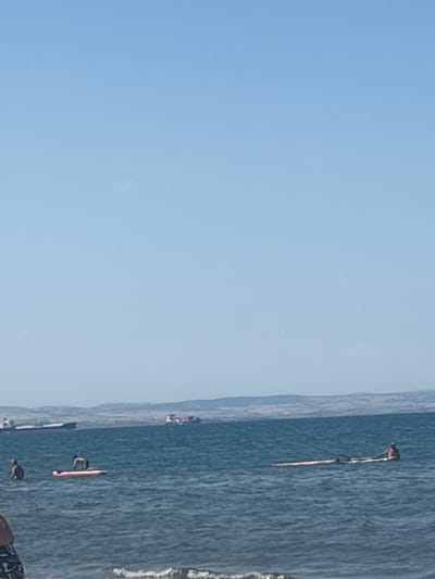 The image shows a body of water under a clear blue sky. In the distance, two cargo ships are visible on the horizon, one to the left and one to the right. A hazy landmass stretches across the background. In the foreground, several people are in the water. One person is swimming, another is on a red paddleboard, and a third is lying on a long paddleboard. The water is calm with gentle waves breaking near the shore in the bottom of the frame. The location is not specified as Atiya, Bulgaria.