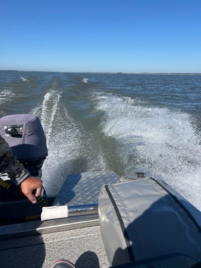 The image shows a view from the stern of a boat moving through choppy water under a clear blue sky. A person's hand, wearing a camouflage-patterned sleeve, is visible in the lower left, resting on part of the boat's interior. To the left, a gray protective cover is on what appears to be an outboard motor. Behind this, white water trails are visible, indicating the boat is in motion. The water extends to a distant horizon line where a faint, low-lying landmass with some buildings can be seen. There are no other discernible features or structures in the immediate environment that would allow for precise location identification within the city of Beregovoe, Russia.