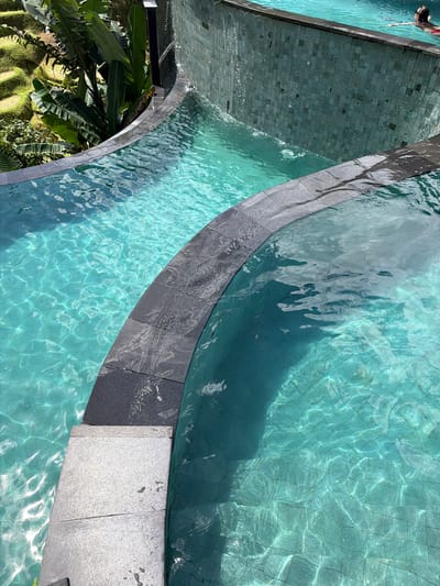 The image depicts a swimming pool with clear, turquoise water located in Tegal Suci, Indonesia. The pool has a tiled bottom and a dark gray tile border. A second pool, also with the same turquoise water, is connected by a cascade. Green tropical plants are visible near one section of the pool. A swimmer is partially visible in the top left corner of the image. Sunlight reflects off the water, creating light patterns on the pool floor.