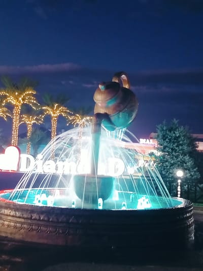 The image captures a decorative fountain at night in Aksukent, Kazakhstan. The fountain features a unique teapot-shaped sculpture from which water cascades into a basin illuminated with bright turquoise lights. Surrounding the fountain are palm trees decorated with numerous small white lights, creating a festive atmosphere. In the background, a building with illuminated signs, including what appears to be "DIAM..." is visible, along with a tall evergreen tree. The sky above is a deep blue, transitioning to darker tones, with subtle clouds suggesting it is late evening or early night. The overall scene depicts a well-lit public space or entertainment area at night.