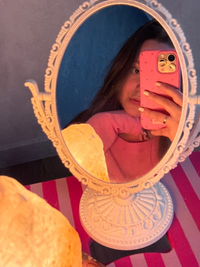 This media file is a selfie taken in a mirror, depicting a person holding a pink phone with a polka dot pattern and a triple camera system. The person is wearing a pink long-sleeved shirt. Their nails are painted with a light color and white polka dots. A ring is visible on their finger. The mirror is ornate, with a white, decorative frame. The reflection shows the person's face, with their eyes looking towards the camera.
The background is a mix of textures and colors. Behind the mirror stand, there are pink and white horizontal stripes, possibly part of a blanket or fabric. To the left of the mirror, a dark blue surface is visible, suggesting a wall or furniture. In the foreground, a large, irregularly shaped object with a rough texture, emitting a warm, orange light, is partially visible. This object appears to be a lamp, possibly a Himalayan salt lamp, given its color and texture.
The lighting suggests an indoor setting, likely during the evening or in a dimly lit room, given the warm glow of the lamp and the overall mood. The image conveys a casual and personal moment, with the selfie being the central activity. There is no visible text in the image. The context of Prikubanskiy, Russia, does not provide specific visual cues within the image itself.