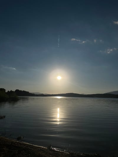 The image shows a body of water, likely a lake or reservoir, with a shoreline visible in the foreground. The sun is setting or rising, casting a bright reflection on the water's surface. Hills and a treeline are silhouetted against the sky in the background. The sky is a gradient of blue, with some scattered clouds. There are no people or man-made structures immediately apparent in the scene, except for what appears to be a vertical text inscription in the upper middle of the sky. The overall scene depicts a tranquil natural landscape at dusk or dawn.