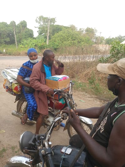 Two people ride a motorcycle loaded with goods. The woman wears a blue patterned dress and headscarf, while the man driving wears a brown jacket. They are stopped on a road beside another man on a motorcycle. He wears a cap and black tank top. The background features trees and vegetation.