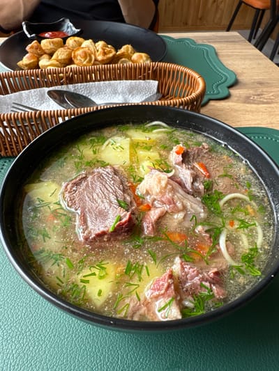 A close-up overhead shot depicts a bowl of meat soup with potatoes, onions, and herbs, likely dill, served on a table. Next to the bowl is a basket containing fried dumplings, a silver spoon, and a small black bowl with red sauce. The bowl is set on a green placemat with a textured surface. The table itself is wooden. The location is likely Semenovka, Kyrgyzstan.