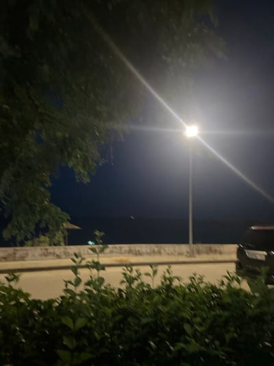 This media file captures a night scene in Nea Karvali, Greece. The primary subjects are a street lamp illuminating the dark sky and surrounding trees. The bright light of the lamp creates strong lens flares that dominate the upper left quadrant of the image, with visible light rays extending outward. To the right, a building's facade, likely made of concrete or stone, is partially visible, with dense green foliage climbing up its side. The foliage is lush and appears healthy.
The scene depicts a quiet night, possibly in a residential area or park, given the presence of trees and a building. The deep blue of the night sky suggests it is well after sunset. There is no indication of specific weather conditions, though the bright light from the lamp suggests clear skies rather than fog or heavy cloud cover.
Notable details include the contrast between the bright artificial light and the dark natural elements of the trees and sky. The way the light interacts with the foliage creates areas of deep shadow and illuminated leaves. In the distance, a faint point of light is visible in the sky, which could be another light source or an aircraft. A small, pointed structure, possibly part of a building or a decorative element, is faintly visible behind the trees on the right side of the frame.
No people are visible in the image, and no specific activity or event is clearly taking place, creating a sense of stillness and quiet. The overall mood is one of peaceful observation of an urban nightscape illuminated by a single strong light source. There is no visible text within the image.