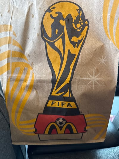 A close-up, eye-level shot shows a McDonald's paper bag with a FIFA World Cup trophy design printed on it. The bag is brown and the design is in yellow and black. The FIFA logo is visible in yellow text on a black background. Below the trophy, there is a red and yellow design that incorporates the McDonald's logo with a soccer ball. The bag is partially in focus, with some areas appearing slightly blurred. The background suggests the bag is inside a vehicle, possibly a car, as indicated by a dark surface and a hint of a blue interior. The lighting is natural, as if taken during the day. There are no people visible in the image. The overall scene is casual and appears to be an everyday setting. There is no information in the image to determine the location in the city of Union City, United States of America.