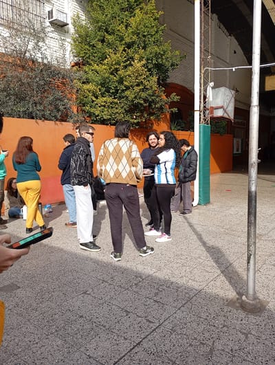 The image depicts an outdoor gathering of approximately ten individuals in a paved courtyard, likely part of a school or community center in La Plata, Argentina, given the architectural style and foliage. The environment features a low, orange brick wall behind the group, a large green tree, and a basketball hoop structure in the background. The pavement is made of grey, speckled tiles.
Several individuals are the focus of the scene. On the left, a person with long brown hair wearing a teal shirt and yellow pants stands facing away from the camera. To their right, a person with dark hair is seated on the ground. A group of four individuals are clustered in the center. One person, wearing a distinctive tan and white argyle sweater, faces away from the camera. Next to them, a person with dark curly hair and a white and blue striped shirt is engaged in conversation. To their left, a man with dark hair and a black jacket is also in conversation. Another man, wearing sunglasses and a black and white patterned jacket, stands to the right of the seated individuals.
Other people are visible in smaller numbers and less detail. Two individuals are standing further back on the right side of the frame. A hand holding a smartphone, with its screen illuminated, is in the lower left corner, suggesting the photo is taken by one of the participants. Objects present include a white thermos and a plastic bottle on the ground near the seated person. The lighting suggests it is daytime, with shadows cast on the pavement.