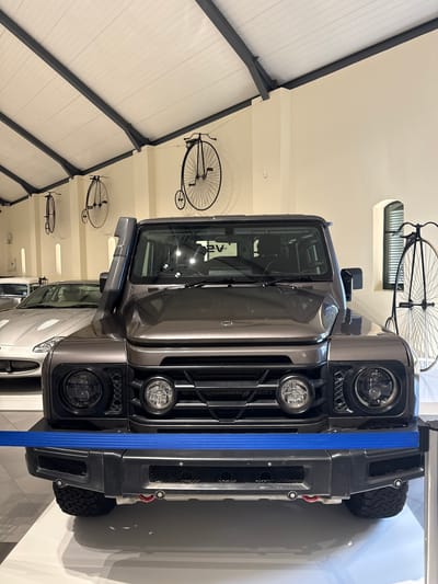 The image shows the front of a dark grey INEOS Grenadier SUV, positioned indoors with a bright blue strap vertically across its front bumper. Round headlights and fog lights are visible, as is the grille. Vintage bicycles are mounted on the wall behind the car. The location is likely within the Stellenbosch Local Municipality, South Africa. The scene appears to be an automotive or design showcase. No people are visible.