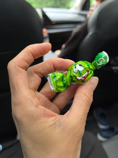 A person's hand holds a green candy wrapper with a cartoon face. The wrapper is crinkled and appears to be made of foil or plastic, with a drawing of a watermelon-like face with large eyes and a wide, smiling mouth showing teeth and a red tongue. The background is blurred, showing the interior of a vehicle with dark seats and some light in the distance. The scene suggests a casual moment, possibly during a car ride. The context of Oktyabr'skii, Russia, is not discernible from the visual elements of the image itself.