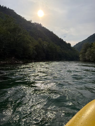 This media file captures a serene moment on a body of water, likely a river, with a lush, tree-covered mountainside on the left and a portion of a yellow kayak on the right. The sun, low in the sky and partially obscured by clouds, casts a warm glow, suggesting it is either early morning or late afternoon. The water's surface is rippled, reflecting the sunlight in a shimmering pattern that creates streaks of brightness. The mountainside is densely forested, indicating a natural, possibly remote, setting. There are no people visible in the frame, and the overall mood is tranquil and peaceful. The location is identified as Zagorice, Bosnia and Herzegovina.
