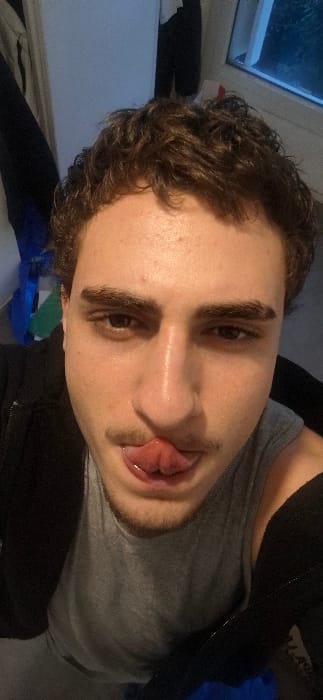 A selfie-style shot of a fair-skinned man with dark curly hair and bushy eyebrows. He is wearing a gray shirt with a black jacket. He is sticking his tongue out. The photo is taken at an odd angle, tilted to the side. The background features a window and some miscellaneous items.